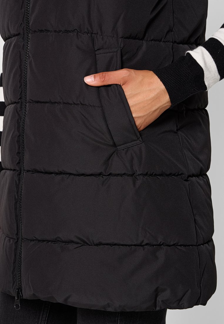 Gilet imbottito nero con cuciture orizzontali, dotato di tasca laterale e chiusura a zip completa, abbinato a maniche a righe in bianco e nero.