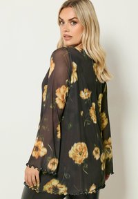 Kimono nero a fiori con grandi motivi di fiori gialli, tessuto trasparente, maniche lunghe con bordi scallop e una vestibilità rilassata.