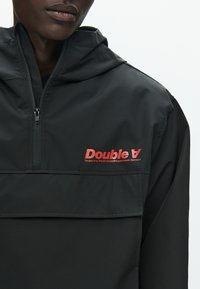 Čierna nepremokavá bunda s polovičným zipsom, kapucňou a červeným logom "Double V" na hrudi. Hladká textúra, minimalistický dizajn.