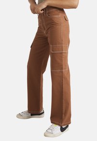 Brune cargo bukser med hvide syninger, der har sidelommer og et lige ben-design. Båret med hvide sneakers.