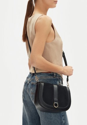 Mujer con top sin mangas beige y vaqueros azules, llevando un bolso cruzado de cuero negro con detalle de anillo dorado.