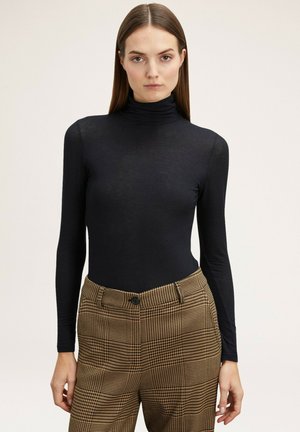 Langärmliger schwarzer Bodysuit aus dehnbarem Stoff mit hohem Kragen, kombiniert mit gemusterten braun karierten Hosen mit Knopfverschluss.