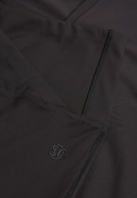 Czarna tkanina sportowa o gładkiej teksturze; posiada kontrastowe stębnowanie oraz mały detal z logo w dolnym rogu.