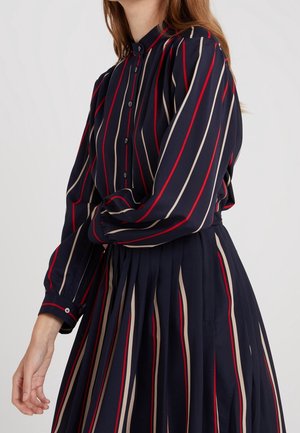 Vrouw draagt een marineblauwe blouse en een geplooide rok met verticale beige en rode strepen, staand tegen een lichte achtergrond.