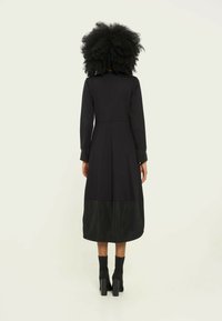 Schwarzes Kleid mit langen Ärmeln, tailliertem Oberteil und einem gepflügten Saum aus einem kontrastierenden Stoff. Matten und glänzenden Texturen zwischen den Schichten.