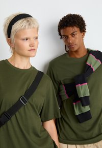 Dwie modelki noszą oliwkowe koszulki. Jedna modelka ma na sobie czarną opaskę na głowę i czarną torbę na ramię. Druga narzuca na siebie pasiasty sweter w czarne, zielone i różowe paski.