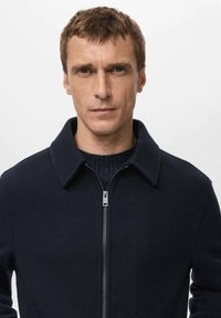 Granatowa kurtka zapinana na zamek z klasycznym kołnierzem, wykonana z gładkiej tkaniny. Posiada detal w postaci dzianinowego kołnierza oraz minimalistyczne elementy metalowe.