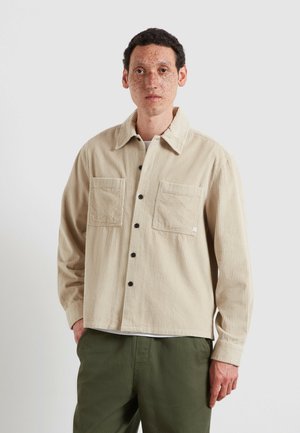 Young man with short curly hair and freckles wearing a beige corduroy button-up shirt and olive green pants against a plain background.