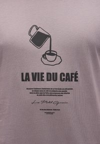 Καφές που ρέει από κανάτα σε φλιτζάνι πάνω σε πιατέλα, με έντονο κείμενο «LA VIE DU CAFÉ» και μικρότερη περιγραφή γαλλικού καφέ από κάτω.