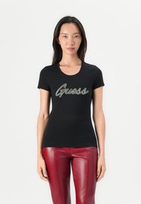 Must have musta T-särk, millel on ümar kaelus, esiküljel on "Guess" metallikult hõbedases kursiivis. Kombineeritud punaste kunstnahast pükstega.