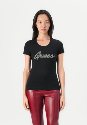T-shirt noir ajusté avec un col rond, portant "Guess" en lettres cursives métalliques argentées. Associé à un pantalon en simili cuir rouge.