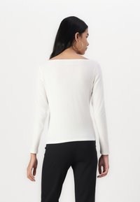 White long-sleeve top with a square neckline. Soft texture and fitted shape. Paired with black pants, showing a clean and simple design.