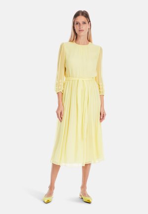 Gelbes, knielanges Kleid mit langen, durchsichtigen Ärmeln und floralen Stickereien an den Bündchen. Mit gebundenem Taillengürtel und fließendem Rock. Getragen mit leuchtend gelben Schuhen.