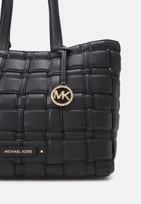 Sac à fourre-tout en cuir noir avec texture tissée, doté de deux bretelles et d'un pendentif en or avec le logo MK. Branding minimal à l'avant.
