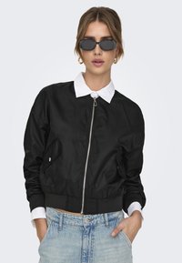 Black bomber jacket with zip front, patch pockets, ribbed cuffs, and hem. Worn over a white collared shirt and paired with black sunglasses.