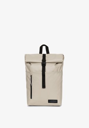 Sac à dos roll-top beige fabriqué à partir d'un matériau lisse et imperméable. Il est doté d'une sangle noire et d'une fermeture éclair, avec un patch logo pour le branding.