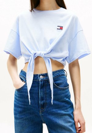 Kobieta ubrana w jasnoniebieski krótki t-shirt zawiązany z przodu, połączony z wysokim stanem niebieskich dżinsów i jedną ręką w kieszeni.