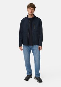 Ung mand står iført en marineblå lynlåsjakke, sort skjorte, lyseblå jeans og sorte vandresko mod en ensfarvet lys baggrund.