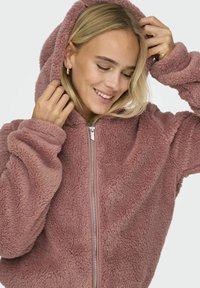 Flauschige rosa Kapuzenjacke mit einem Reißverschluss an der Vorderseite, gerippten Bündchen und einem plüschigen, strukturierten Stoff. Das Design umfasst eine lockere Passform und eine große Kapuze.
