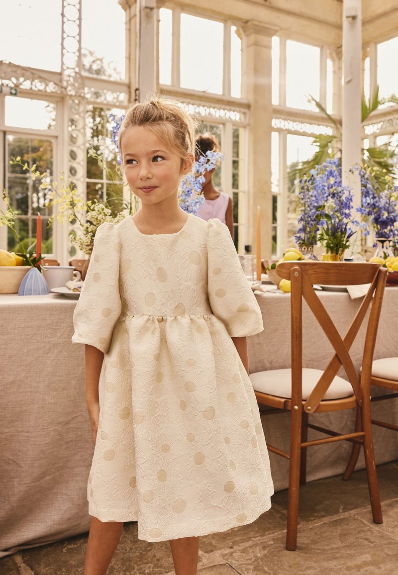 Jeune fille en robe crème à manches bouffantes avec motifs floraux, debout près d'une table à manger décorée de fleurs et de bougies.