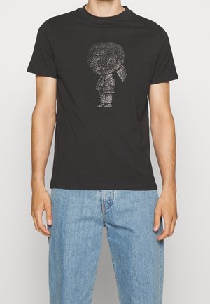 Persoon draagt een zwart T-shirt met een glinsterend zilveren afbeelding van een cartoonfiguur en lichtblauwe spijkerbroek tegen een effen achtergrond.