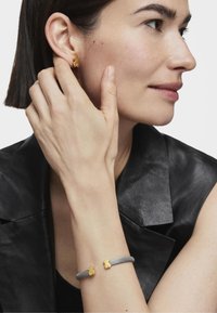 Pendientes en forma de oso de oro y una pulsera de malla de plata con acentos de oso de oro. El modelo lleva una blusa de cuero negra y tiene una piel suave.