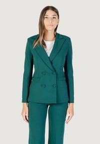 Una modelo viste un blazer de doble botonadura en color teal y pantalones a juego, de pie contra un fondo claro con una expresión neutral.