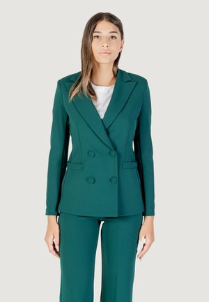 Una modella indossa un blazer doppiopetto color teal e pantaloni abbinati, in piedi su uno sfondo chiaro con un'espressione neutra.