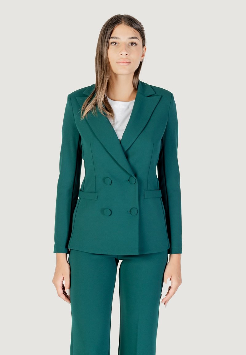 Una modelo viste un blazer de doble botonadura en color teal y pantalones a juego, de pie contra un fondo claro con una expresión neutral.