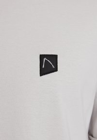 Bílá bavlněná tričko s malou černou trojúhelníkovou nášivkou s bílými kresbami. Hladký povrch, minimalistický design.