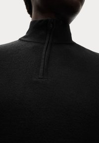 Schwarzer Strickpullover mit hohem Kragen und halbem Reißverschluss, der aus einem strukturierten Stoff besteht und einen glatten Abschluss am Kragen hat.