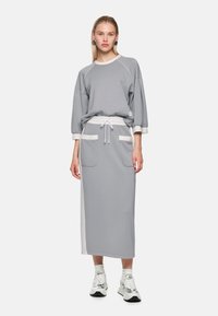 Graues Trikotkleid im übergroßen Sweatshirt-Stil mit weißen Akzentdetails, Seitentaschen und Taillenschnur, kombiniert mit Sneakern.