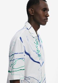 Camicia bianca a maniche corte con motivi astratti blu e verdi. Presenta un colletto piatto e una texture morbida con sottili righe.