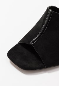 Svart mocka öppen tå mule-sko med fyrkantig tå och synlig vit stickning längs den främre överkanten.