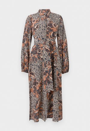 Frau trägt ein langärmliges, knöchellanges Kleid mit blauen, orangefarbenen und weißen Blumen- und abstrakten Mustern und steht mit einer Hand in der Tasche.