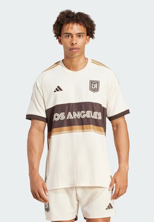 Maglia in jersey crema con una banda orizzontale marrone scuro che presenta "LOS ANGELES" in bianco. Dettagli in oro e logo Adidas sulla spalla. Texture liscia.