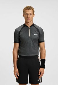 Koszulka sportowa z krótkim rękawem w ciemnoszarym kolorze z czarnymi akcentami, z zamkiem przy kołnierzu i logo. W połączeniu z czarnymi szortami i pasującą opaską na nadgarstek.