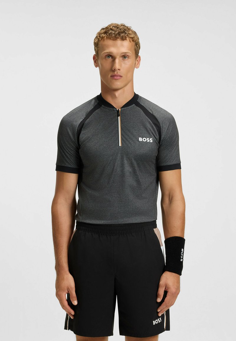 Koszulka sportowa z krótkim rękawem w ciemnoszarym kolorze z czarnymi akcentami, z zamkiem przy kołnierzu i logo. W połączeniu z czarnymi szortami i pasującą opaską na nadgarstek.