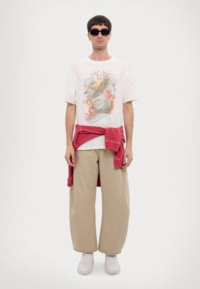 Camiseta blanca de gran tamaño con gráfico de águila y llamas, combinada con pantalones de pierna ancha beige y un suéter rojo atado a la cintura. Zapatillas blancas.