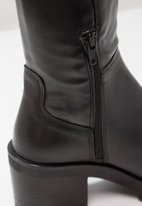 Gros plan sur une botte cheville en cuir noir avec fermeture éclair latérale et petit talon empilé sur fond blanc.