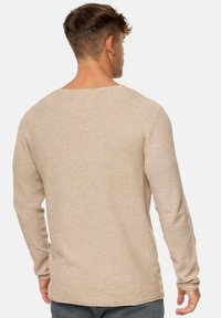 Beige, pitkähihainen neule, jossa on teksturoitu neulos, leveä pääntie ja hieman kaareva helmakaari takana.