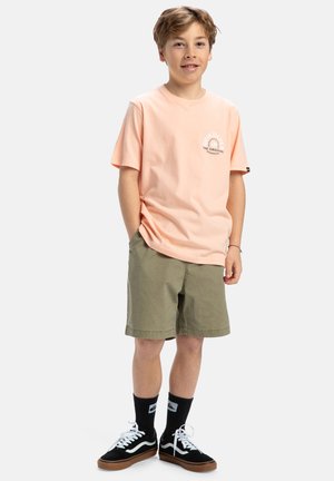 Camiseta de algodón rosa claro con un logo circular, combinada con pantalones cortos verde oliva. Zapatillas negras con acentos blancos y calcetines oscuros completan el atuendo.
