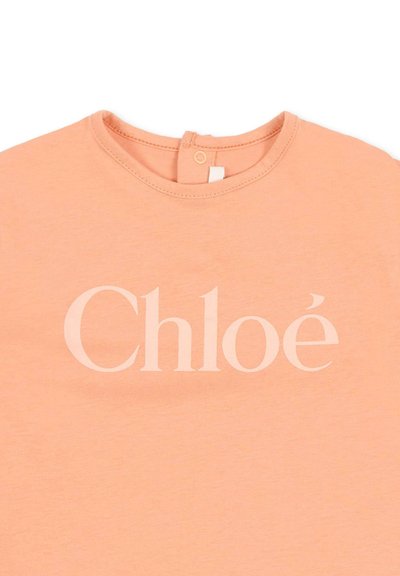 Brzoskwiniowa koszulka dla malucha z okrągłym dekoltem i zapięciem na guziki z tyłu, z dużym napisem "Chloé" w białych literach na przodzie.