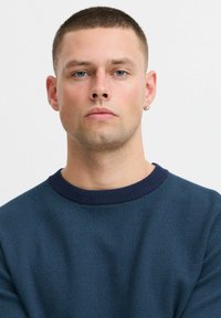 Temno modra pletena jopica z teksturiranim vzorcem in rebrastim mornarsko modrim ovratnikom. Model ima kratke, svetle lase in uhano.