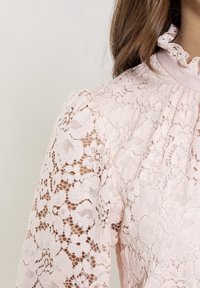 Krajková blůza v jemné růžové barvě. Vyznačuje se složitými květinovými krajkovými vzory a volánovým límcem. Průhledné dlouhé rukávy umocňují křehký design.
