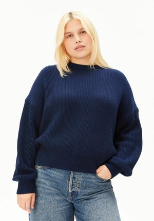 Jeune femme aux cheveux blonds raides portant un pull côtelé bleu marine et un jean bleu, debout avec une main dans la poche.