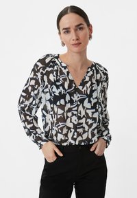 Blouse in zwart, wit en blauw abstracte print; lichtgewicht, doorschijnende stof; V-hals; lange mouwen, plooien bij de schouders; gecombineerd met zwarte broek.