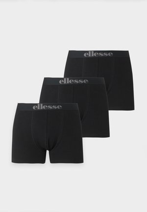 Hombre sin camiseta, de pie descalzo, vistiendo calzoncillos bóxer negros con cinturilla con logo, posando frente a un fondo blanco liso.
