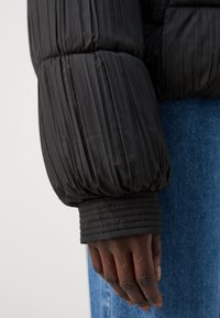 Mâneca unei jachete puf negre cu pliseuri verticale, cu manșete elastice și textură netedă. Mână sprijinită pe un material din denim albastru.