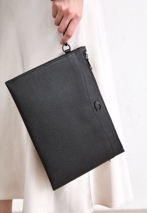 Hand die een zwarte leren clutch met rits vasthoudt tegen een witte rok of jurk als achtergrond.